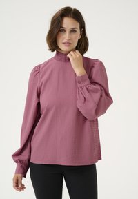 Blouse rose en tissu texturé, avec un col haut froncé, des manches longues bouffantes et des poignets boutonnés, associée à un pantalon noir.