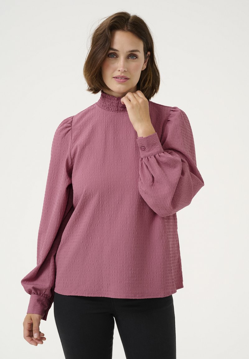 Blouse rose en tissu texturé, avec un col haut froncé, des manches longues bouffantes et des poignets boutonnés, associée à un pantalon noir.