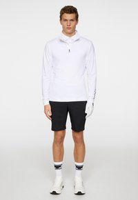 Valkoinen pitkähihaisuus paita, jossa on neljänneksen vetoketju, yhdistetty mustiin shortseihin. Mallilla on päällä valkoiset hanskat ja sukat, joiden kuviointi on musta-valkoinen raita.