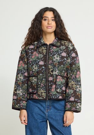 Femme aux longs cheveux bouclés portant une veste zippée à motif floral et un jean bleu, debout devant un fond uni.