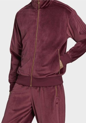 Sportowy dres z aksamitu w kolorze bordo z złotymi detalami na zamkach, ściągaczami na rękawach i subtelnym haftowanym logo na kurtce i spodniach.