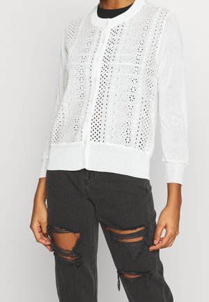 Hvid knap-op cardigan med perforerede mønstre, båret over sort revnede jeans, vist fra nakke til midt på låret.