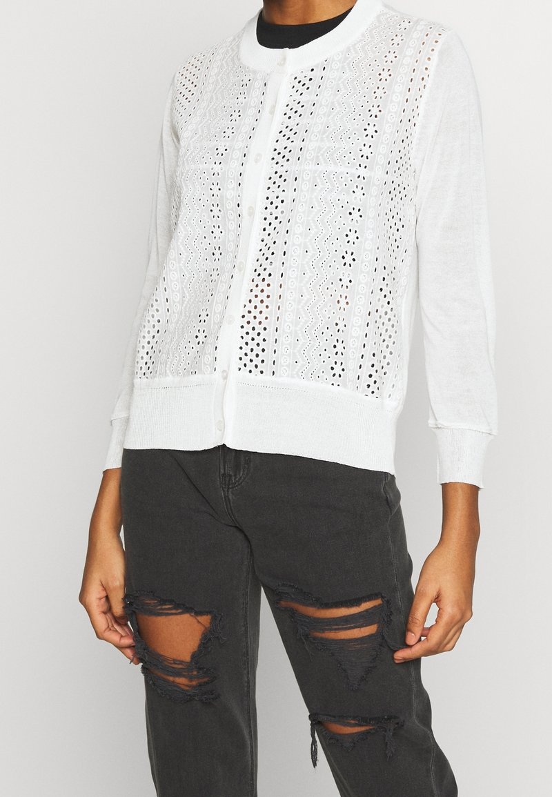 Cardigan blanc boutonné avec des motifs perforés porté sur un jean noir déchiré, montré du cou à la mi-cuisse.