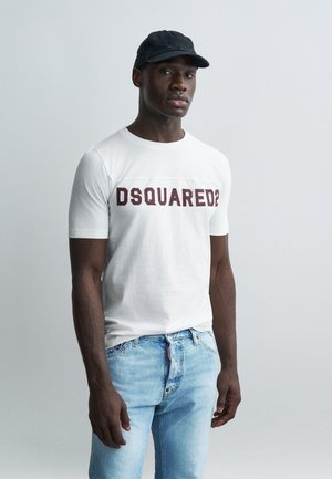 Homme portant une casquette noire, un t-shirt blanc DSQUARED2 et un jean bleu clair, debout devant un fond uni gris clair.