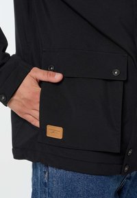 Persona che indossa una giacca nera con una mano dentro una tasca frontale con patta che ha bottoni a scatto e una piccola toppa con logo marrone.