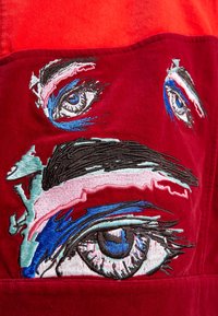 Gros plan sur un tissu rouge avec un motif abstrait brodé présentant trois yeux bleus et noirs détaillés avec des accents colorés.