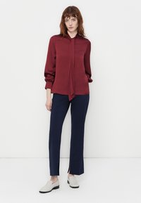 Blusa bordeaux con un laccio, maniche lunghe con polsini elasticizzati, abbinata a pantaloni a quadri blu navy e scarpe slip-on di colore chiaro.