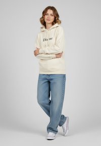 Felpa beige con la scritta "Divine" in nero, dotata di cappuccio con cordino. Abbinata a jeans a gamba larga di colore blu chiaro e sneaker bianche.