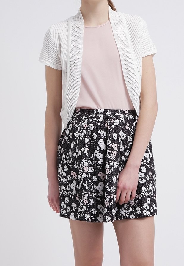Femme portant un cardigan en maille blanc à manches courtes, un haut rose pâle et une jupe noire avec un motif floral blanc et rose clair.