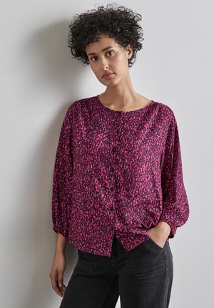 Losse, langwerpige blouse met lange mouwen in een levendige roze kleur met een zwart abstract patroon, voorzien van een ronde halslijn en knoopdetails. Gepaard met zwarte broek.