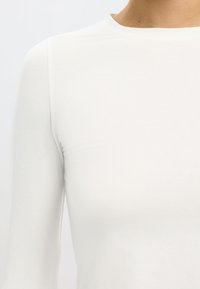 Valkoinen pitkähihaiset paita, joka on valmistettu sileästä, joustavasta kankaasta. Ominaisuuksiin kuuluu korkea kaula-aukko ja vartalon sekä hartioiden tiukka muotoilu.