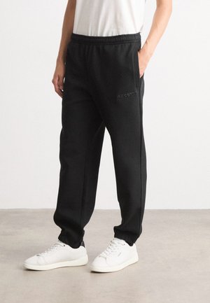 Pantalons de survêtement noirs en matériau doux avec une taille élastique et des poches latérales ; associés à des baskets blanches. Design minimaliste.
