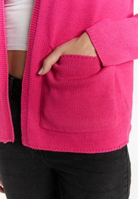 Pink knitted cardigan with a textured finish, featuring a front pocket and ribbed cuffs. The bottom edge is slightly fringed.