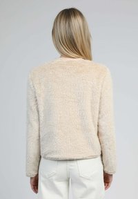 Personne blonde portant un haut à manches longues en crème duveteux et un pantalon blanc, de dos contre un fond gris uni.