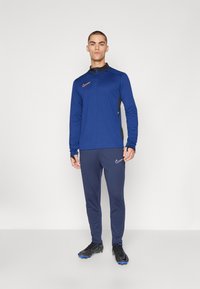 Tengerészkék sportfelső cipzáros nyakkal, fekete részletekkel és narancssárga Nike logóval; tengerészkék nadrággal és fekete sportcipővel, kék talppal párosítva.