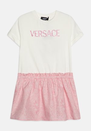 Valkoinen lyhythihainen mekko, jossa on vaaleanpunainen kukkakuvioitu, rypytetty helma ja "VERSACE"-logo vaaleanpunaisilla paljeteilla rinnassa.