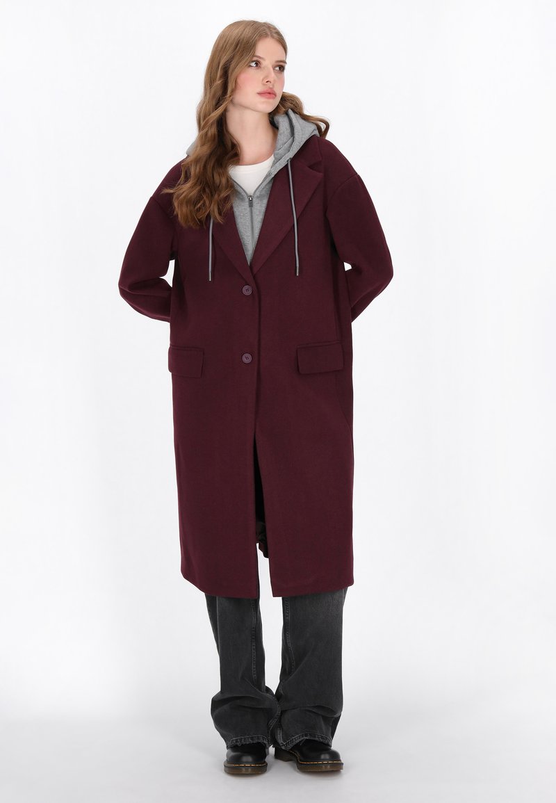 Cappotto lungo bordeaux con un design strutturato, dotato di due tasche frontali, chiusura con un solo bottone e una felpa con cappuccio grigia sotto.