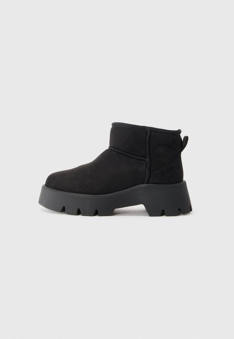 Bottine noire avec semelle épaisse à crampons, tige en suédine douce, détails de couture et languette au talon sur fond gris clair uni.