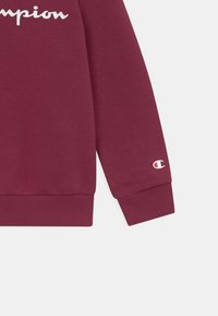 Felpa borgogna con polsini a costine, con il testo bianco del logo "Champion" sul petto e un piccolo logo sulla manica. Tessuto morbido.