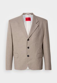 Blazer beige su misura con spalle strutturate, design monopetto, tre bottoni neri, due tasche frontali e etichetta HUGO in rosso.