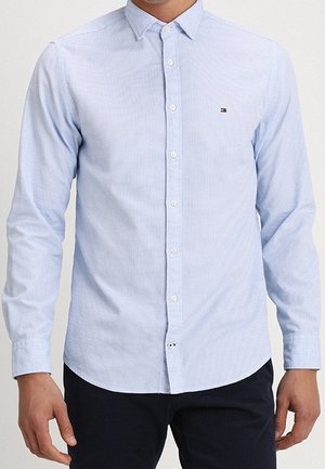 Mężczyzna noszący jasnoniebieską, długim rękawem, koszulę na guziki z subtelnym wzorem i małym logo na piersi, w połączeniu z ciemnymi spodniami.