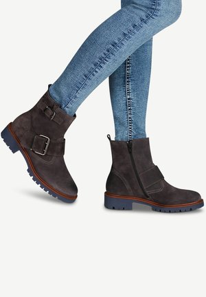 Botas de ante negras hasta el tobillo con cremallera lateral y una correa con hebilla, suela marrón en contraste y suelas de goma texturizadas. Jeans de mezclilla visibles.
