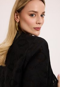 Czarna, teksturowana odzież o subtelnym wzorze, luźny krój. Duże, złote kolczyki w kształcie kół podkreślają całość. Modelka ma proste, blond włosy.