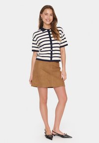 Maglione a righe con maniche corte in navy e crema, abbinato a una mini gonna in suede beige. La donna indossa ballerine nere a punta e mostra i capelli naturali.