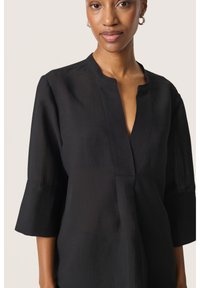 Svart blus med V-ringning, trekvartslånga klockärmar, texturerad tyg och subtila vertikala ränder. Har en minimalistisk design.