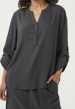 Vrouw draagt een donkergrijze blouse met lange mouwen, twee knopen en opgerolde mouwen, hand rustend in de zak van bijpassende broek.