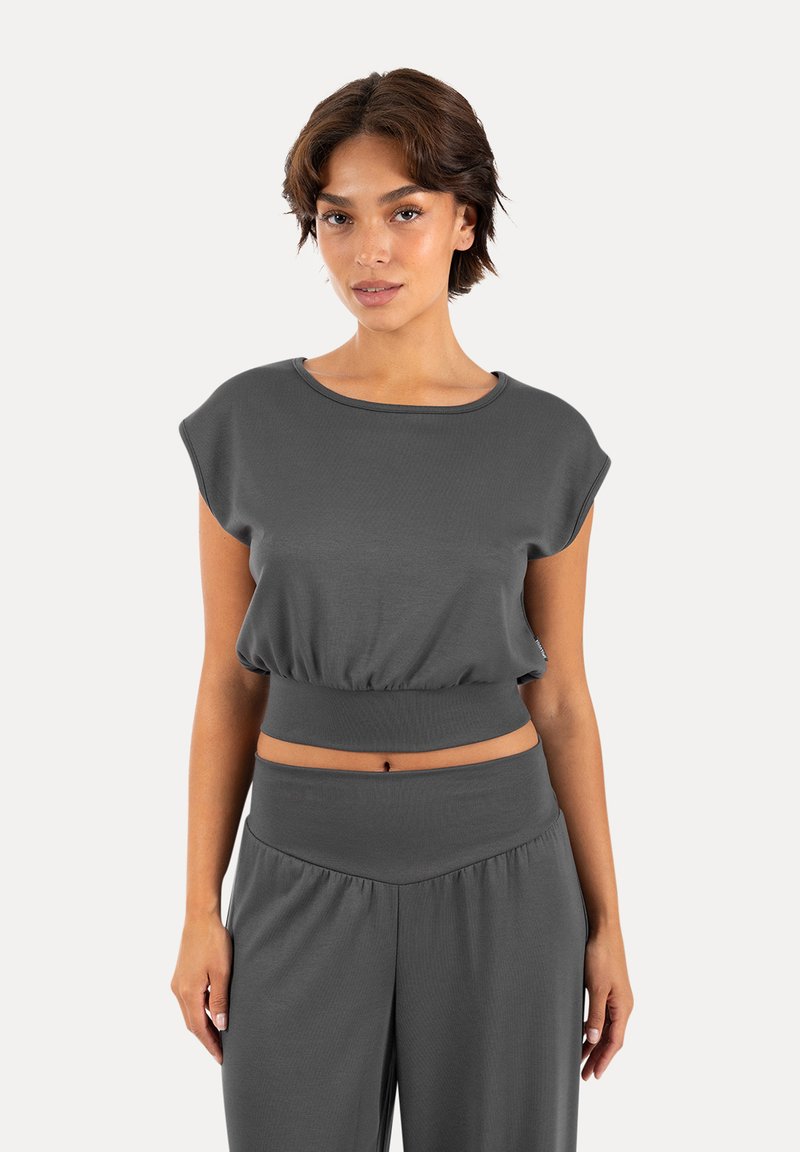 Jeune femme aux cheveux courts et bruns portant un crop top gris foncé à manches courtes et un pantalon taille haute assorti, debout sur fond blanc.
