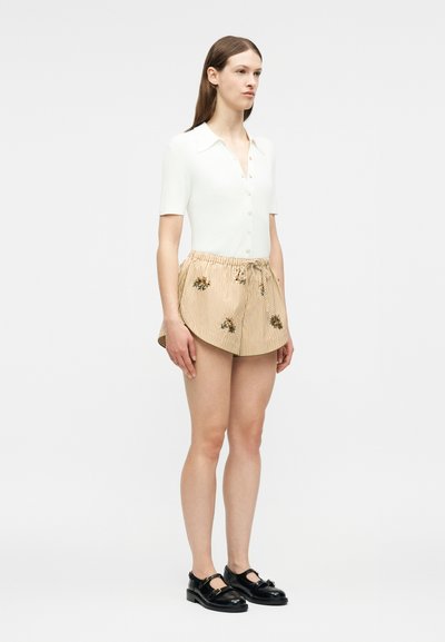 Γυναίκα που στέκεται, φοράει λευκό κοντομάνικο πουκάμισο με κουμπιά, μπεζ ριγέ σορτς με λουλουδένιο σχέδιο και μαύρα παπούτσια Mary Jane.