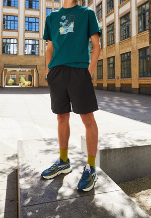 Mand står udendørs iført sorte shorts, grøn t-shirt med grafik, olivengrønne sokker og blå-hvide sneakers foran en murstensbygning.