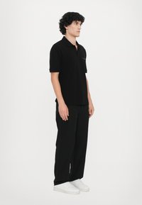 Svart polo-skjorte med korte ermer, myk tekstur, klassisk krage og diskret logodetalj, kombinert med sorte bukser og hvite joggesko.