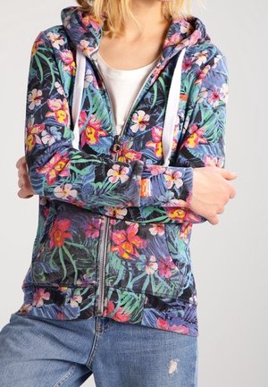 Personne portant un sweat à capuche coloré à fleurs avec une fermeture éclair, des cordons blancs et un jean bleu avec une petite déchirure sur la cuisse.