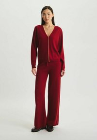 Ensemble en tricot rouge comprenant un cardigan à col en V avec fermeture éclair et poches, associé à un pantalon large, le tout en tissu doux et texturé.
