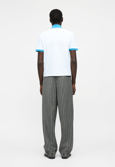Άνδρας που στέκεται με την πλάτη προς τα εμπρός, φορώντας λευκό πουκάμισο με κοντά μανίκια, μπλε γιακά και μανσέτες, και γκρι ριγέ παντελόνι με φαρδιά μπατζάκια.