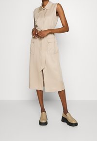 Beżowa sukienka bez rękawów z przodem na guziki, dwoma przednimi kieszeniami, talią z paskiem i długością do kolan. Do tego beżowe wsuwane buty.