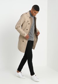 Beige trenchcoat med store knapper, med notched krage og knelængde design. Båret over en grå sweater og parret med sorte bukser og hvide sneakers.