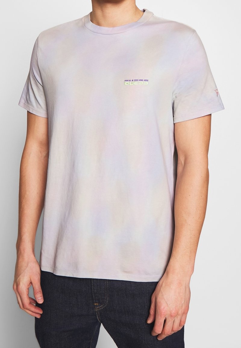 T-shirt violet clair en coton doux, doté d'un motif tie-dye subtil et d'un logo "GUESS" réfléchissant sur la poitrine. Manches courtes.