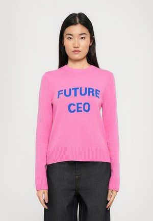 Kvinne med langt mørkt hår iført en rosa genser med blå tekst "FUTURE CEO" og mørke bukser, stående mot en enkel lys bakgrunn.