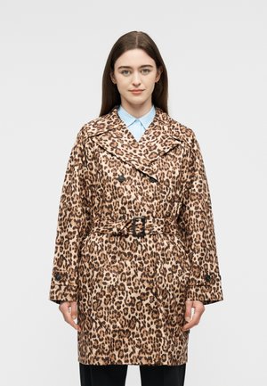 Femme aux longs cheveux bruns portant un manteau à double boutonnage imprimé léopard avec une ceinture, debout devant un fond clair uni.