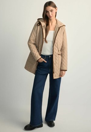 Beżowa pikowana kurtka z kapturem, brązowymi akcentami, dwoma bocznymi kieszeniami oraz dopasowanym w talii krojem, zestawiona z szerokimi, niebieskimi dżinsami i czarnymi butami.