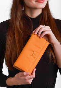 Oranžová kabelka z látky s hladkým povrchem, skládací design a detail zigzagového stehu. Drží ji osoba oblečená v černém topu.