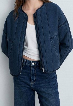 Person iført en mørkeblå quiltet jakke over en hvid cropped tanktop og højtaljede mørkeblå jeans mod en lys baggrund.