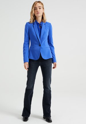 Mėlynas pritaikytas švarkas su viena sagte, iškiliais apykaklėmis ir dviem priekiniais kišenėmis, derinamas su tamsiomis platėjančiomis džinsais ir juodais kulkšnies botais.