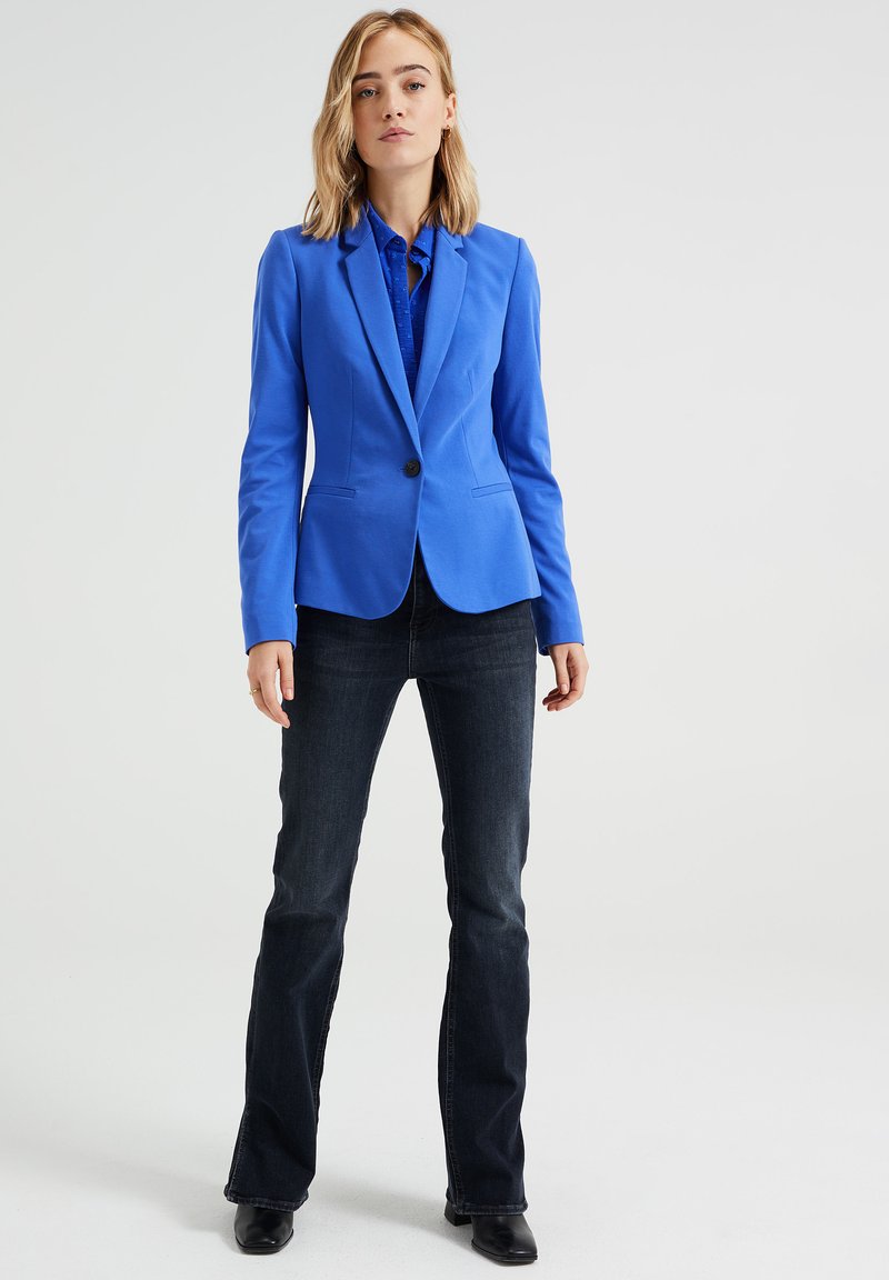 Blauer maßgeschneiderter Blazer mit einem einzelnen Knopf, Reverskragen und zwei vorderen Taschen, kombiniert mit dunklen Schlagjeans und schwarzen Ankle Boots.