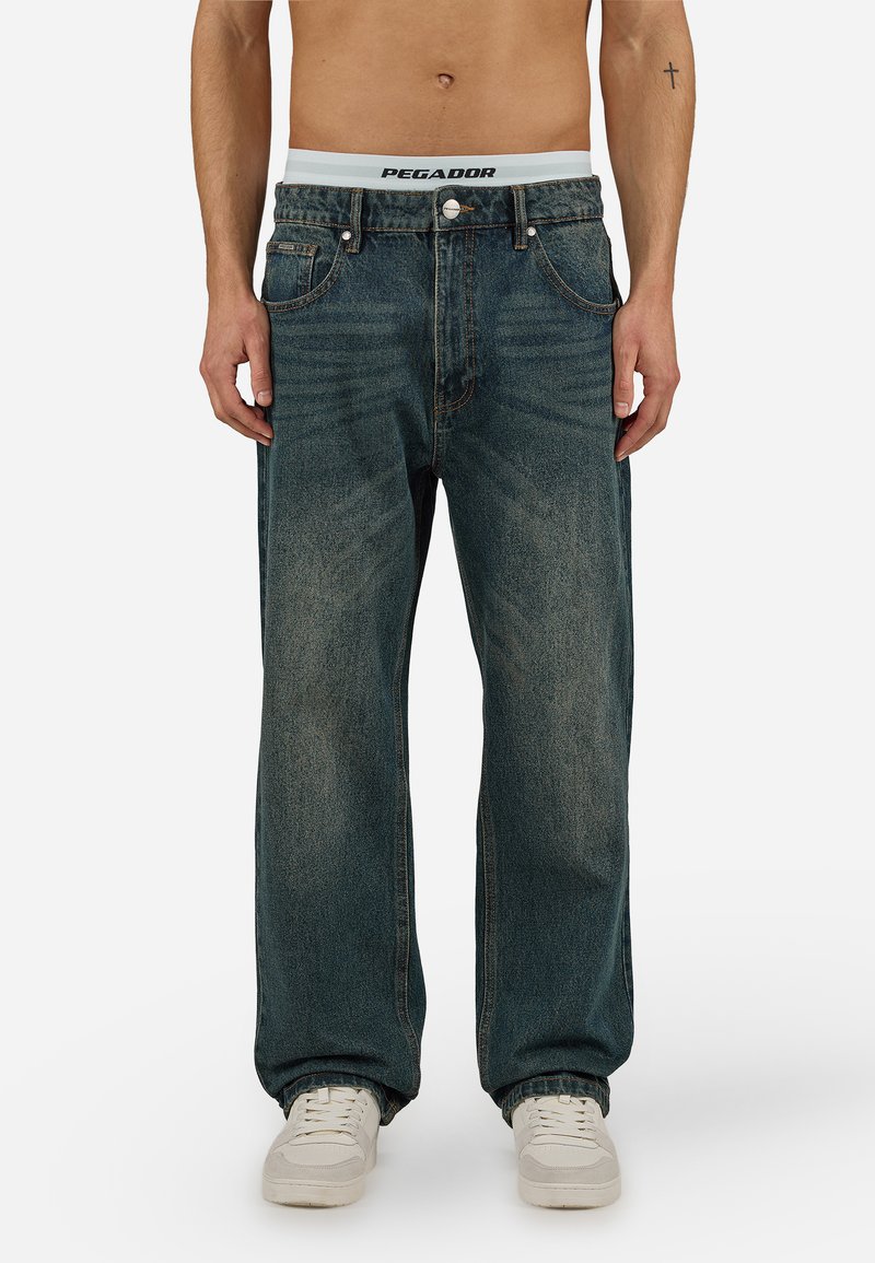 Άνδρας που φοράει φαρδιά ξεβαμμένα μπλε τζιν, λευκά αθλητικά παπούτσια και λευκό εσώρουχο με ρίγα μέσης που φέρει το λογότυπο "PEGADOR", στέκεται μπροστά σε απλό φόντο.