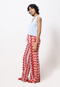 Hellblaues, ärmelloses Oberteil kombiniert mit bunten, gemusterten weit geschnittenen Hosen in Rot-, Pink- und geometrischen Formen. Rote Schuhe runden das Outfit ab.