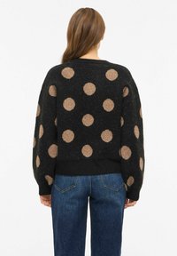 Vabaajakampsun, millel on must alus ja suured beežid täpid, valmistatud tekstureeritud kudumiskangast ning mille lõige on mugavalt lõdvestunud.